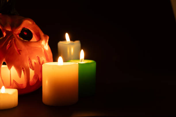 A hollow Jack-O'-Lantern, lit by inner candles, gazes eerily. Surrounding colored candles are placed on the left, all against a dark, black background in this horizontal photo.