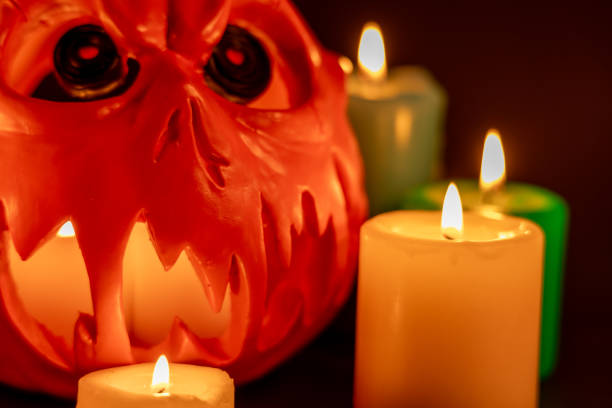 A detailed horizontal photo of a hollow Halloween pumpkin with a piercing stare. It has candles inside and colorful candles around it on a black background