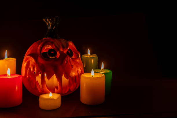 Side view of a hollow Halloween pumpkin with candles inside and colored candles around it. Positioned to the left on a black background, creating a spooky atmosphere.