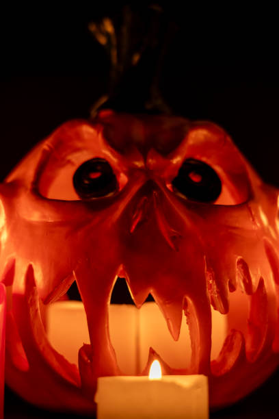 Vertical image featuring a hollow pumpkin with an intense gaze, candles glowing inside, and colorful candles around, all set against a black background.