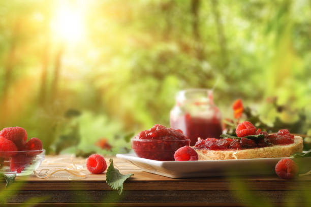 Homemade raspberry jam in glass jars and slice of bread on wooden table and nature background. Front view.