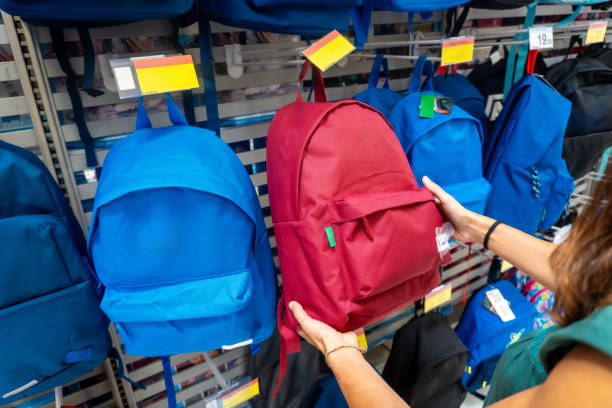 A woman is holding a red backpack in a store. There are many backpacks on display, including blue and green ones. Back to school concept