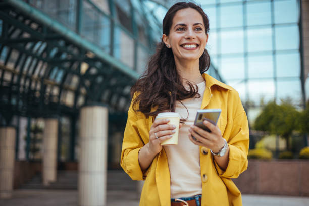 istock Smiling Woman Holding Coffee and Smartphone Outdoors in Urban Setting 2170478430