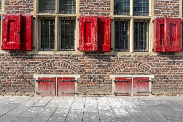 A historic brick building with red wooden shutters and large windows. A small balcony with pink flowers adds charm. The stepped gable and pulley beam hint at Dutch architectural style.