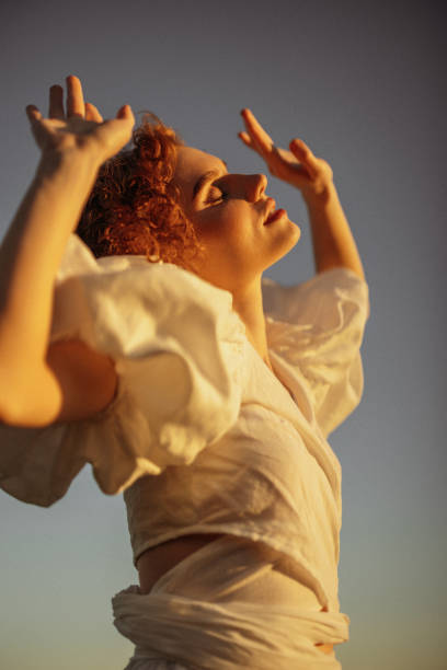 This evocative image depicts a young woman with curly red hair walking through a sunlit meadow during late afternoon. Her vintage-style white dress, with puffed sleeves and a tie at the neck, adds a timeless elegance to the scene. She appears absorbed in the moment, gently touching the wildflowers as she walks by. The warm, natural lighting and the open field background enhance the pastoral and peaceful ambiance, evoking a sense of freedom and connection to nature.