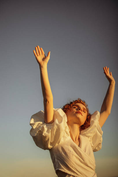 This evocative image depicts a young woman with curly red hair walking through a sunlit meadow during late afternoon. Her vintage-style white dress, with puffed sleeves and a tie at the neck, adds a timeless elegance to the scene. She appears absorbed in the moment, gently touching the wildflowers as she walks by. The warm, natural lighting and the open field background enhance the pastoral and peaceful ambiance, evoking a sense of freedom and connection to nature.