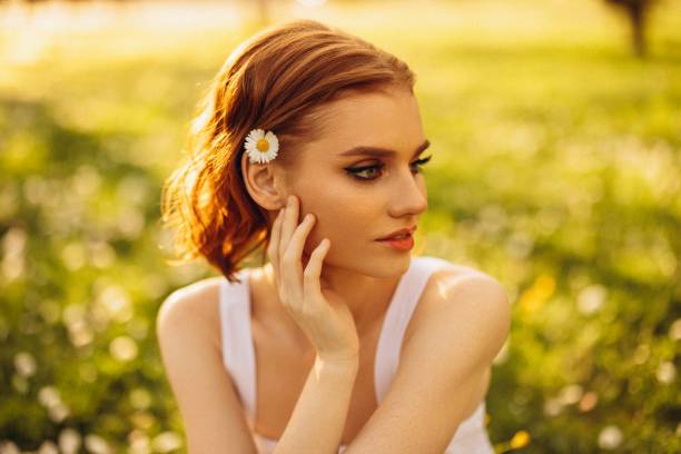 This captivating portrait captures the gentle grace of a young woman lost in contemplation amidst a backdrop of soft, natural light. Her subtle, reflective pose blends beautifully with the warm, golden hues of the surrounding landscape, creating a peaceful and introspective atmosphere. The soft focus on the lush greenery enhances the dreamlike quality of the scene, highlighting the woman's serene expression and delicate features.