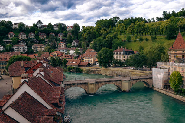A picturesque view captures a historic bridge over a river in Bern, Switzerland, framed by vibrant greenery and traditional Swiss buildings on the banks.