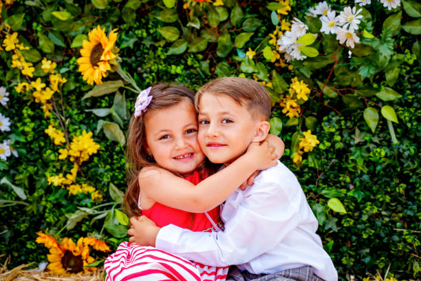 Two children embracing in the garden on a sunny summer day. The outdoor floral decorations in the background suggest a special celebration. One of the children is wearing a white shirt, the other a red and white sleeveless dress. Both children are smiling warmly, radiating a feeling of love and closeness. The photo beautifully depicts the innocence of childhood, friendship and the joy of moments shared in a cozy, festive setting.