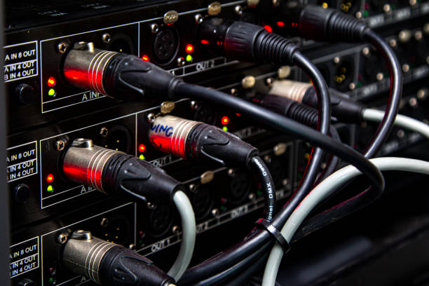 A detailed view of an audio patch panel installed in a recording studio wall, with a 1/4 inch instrument cable plugged into one of the jacks. The wooden floor and patterned carpet add to the warm and vintage atmosphere of the space. Ideal for themes related to music production, recording environments, or audio equipment.