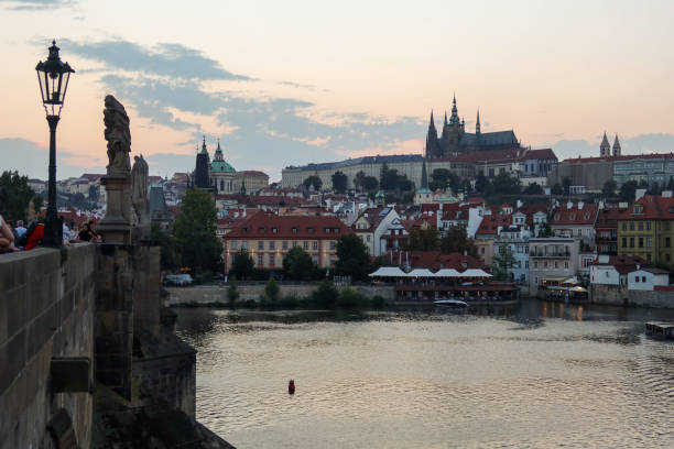 A twilight view of Prague Castle from Charles Bridge, with sunset colors reflecting on the calm Vltava River. Silhouetted towers rise against the glowing sky, creating a serene, romantic atmosphere.