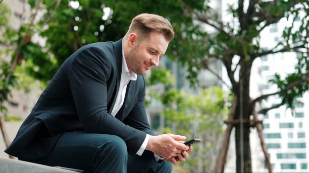 istock Businessman sitting at park while working on phone by reading report. Urbane 2170544156