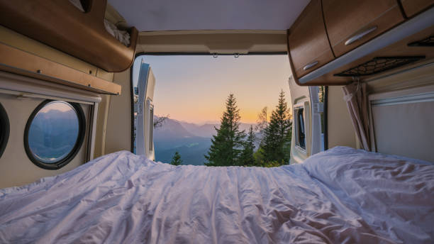Inside a camper van, warm bedding offers comfort as the sunrise unveils breathtaking mountains outside. Lush trees frame the view, inviting adventure in the tranquil Austrian landscape.