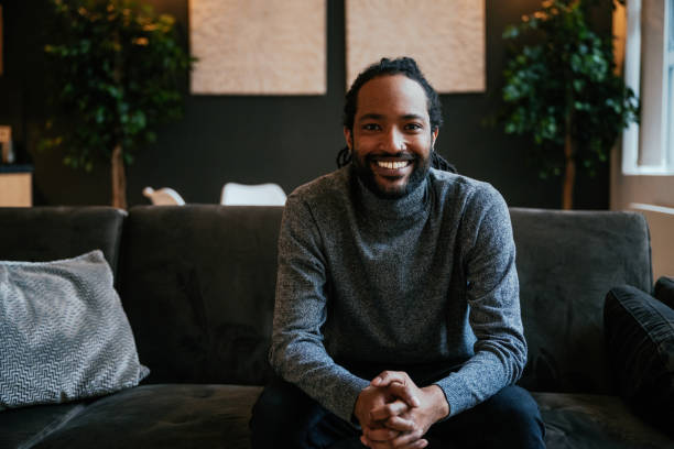 istock Smiling black man with a beard sitting on a dark sofa with hands clasped, surrounded by indoor plants and soft lighting. 2170575002
