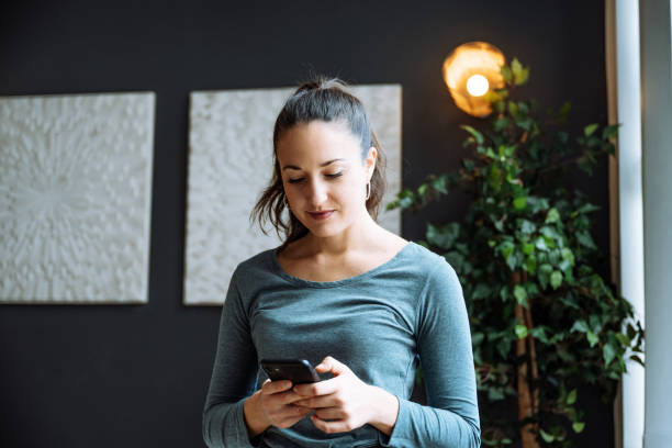 istock Young woman with ponytail using smartphone while standing in a stylish living room with modern decor and green plant. 2170575132