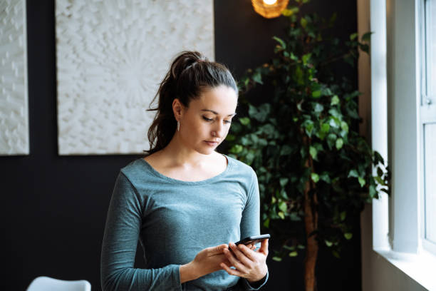 istock Serious woman with ponytail using smartphone while standing in a stylish living room with modern decor and green plant. 2170575505