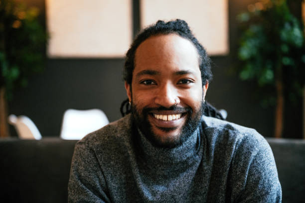 istock Smiling black man with a beard sitting on a dark sofa with hands clasped, surrounded by indoor plants and soft lighting. 2170575508