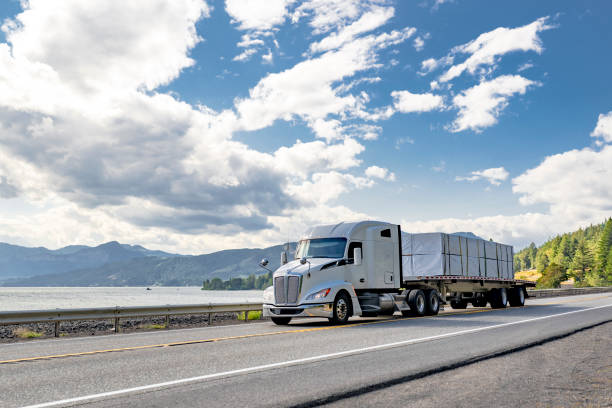 istock Bonnet powerful white big rig semi truck transporting covered fastened cargo on flat bed semi trailer running on the road along the railroad and river 2170582176