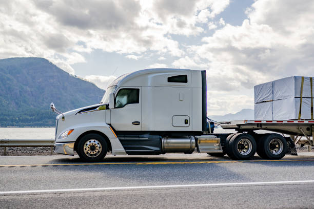 istock Big rig white semi truck with aerodynamic profile transporting fastened cargo on flat bed semi trailer driving on the highway road along the river 2170582420