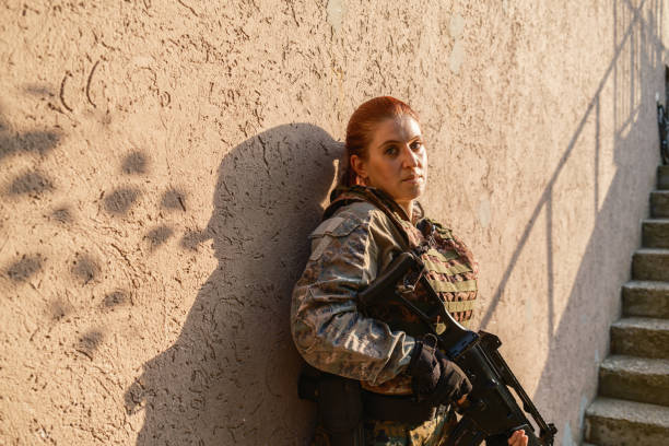 Military training: armed female  soldier in camouflage gear taking a break armed with with rifle against the outdoor wall, during practising military discipline and teamwork.