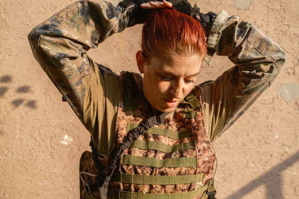 Military training: armed female  soldier in camouflage gear taking a break armed with with rifle against the outdoor wall, during practising military discipline and teamwork.