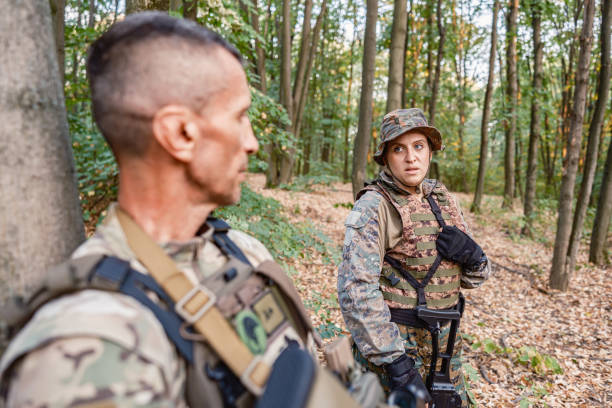 Military training: two soldiers- male and female, in camouflage gear taking a break with rifles in wooded area during military training, practising military discipline and teamwork during outdoor operation.