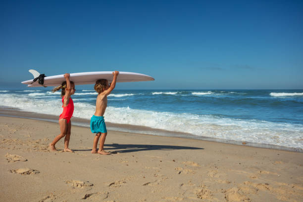 Beach-bound kids team up to carry a surfboard with the expansive ocean and clear skies behind them, surf school concept