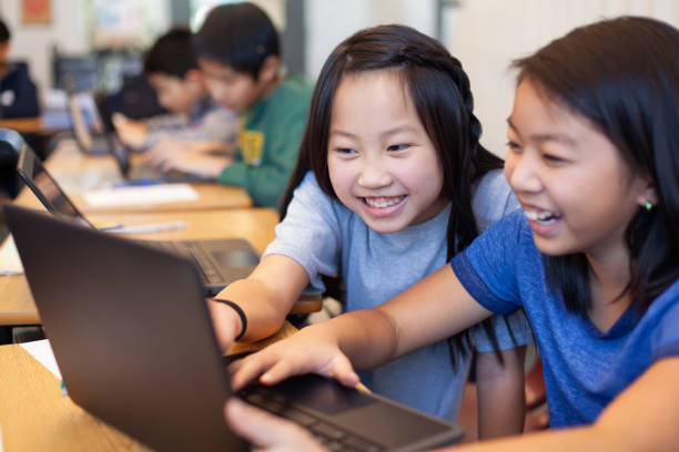 Two Asian Chinese girls students collaborate together using laptop computer in elementary school classroom