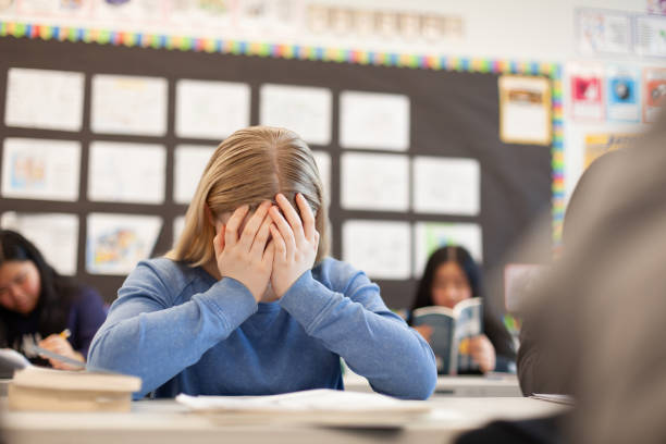 istock Elementary school junior high girl student sitting at desk in classroom with hands on her face in frustration sadness anxiety worried 2170616238