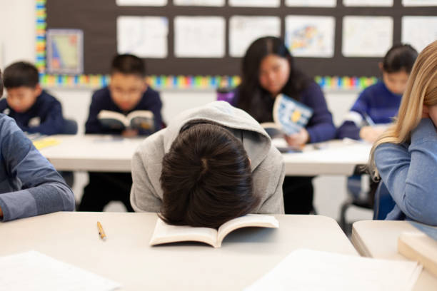 Elementary school junior high boy student sitting at desk in classroom with head in book in frustration sleeping tired exhaustion boredom