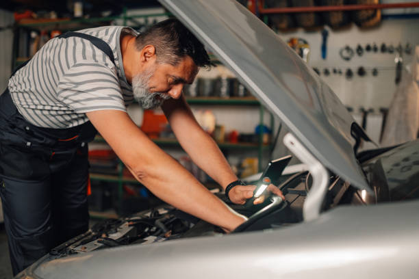 istock Side view of auto mechanic with lamp looking under car hood at workshop 2170631549