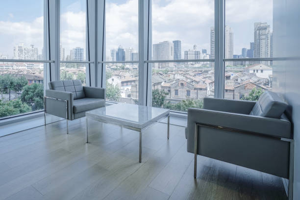 A bright and spacious modern office lounge area with floor-to-ceiling windows offering panoramic views of an urban skyline. The room features minimalistic furniture, including sleek, grey leather armchairs and a matching marble coffee table. Natural light floods the space, emphasizing the clean lines and contemporary design, while the expansive windows create an airy, open atmosphere. The contrast between the bustling cityscape and the serene interior makes this a perfect place for relaxation or casual meetings in a professional setting.