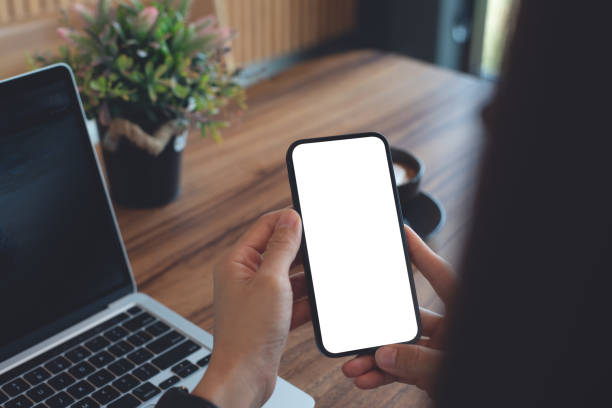 istock mockup blank white screen cell phone for web template or mobile app design. Woman hand using mobile phone during working on laptop computer on wooden table at coffee shop or home office 2170640244