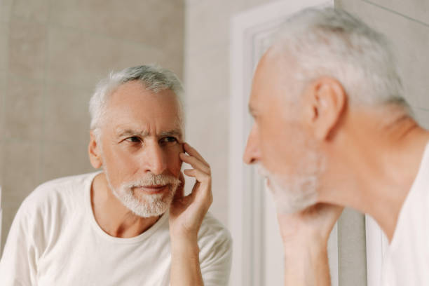 Elderly, bearded man touching face checking wrinkles in mirror, skin care