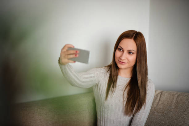 A young woman with straight brown hair is sitting on a couch, holding her smartphone and taking a selfie. She is dressed in a light sweater and appears relaxed and confident in a cozy indoor setting.