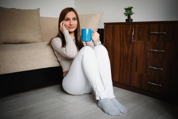 A young woman with brown hair is sitting on the floor, leaning against a couch while talking on the phone and holding a blue coffee mug. She is dressed casually in a light sweater and white jeans, enjoying a relaxed moment at home.