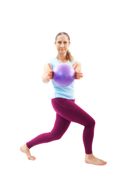 Focused woman in a lunge position holding a Pilates ball extended forward, emphasizing balance, core strength, and flexibility, isolated on a white background.