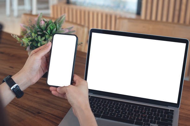 istock Mockup blank white screen cell phone and laptop for web template or mobile app design. Woman hand using mobile phone during working on laptop computer on wooden table at coffee shop or home office 2170662427
