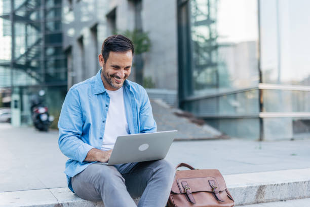 istock A handsome, cheerful, smiling man sitting on steps and using a laptop, with modern buildings in the background. 2170679963