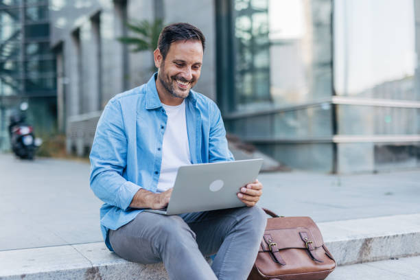 istock A handsome, cheerful, smiling man sitting on steps and using a laptop, with modern buildings in the background. 2170679965