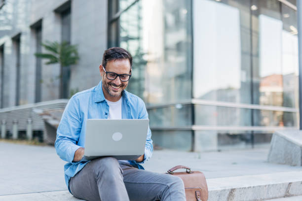 istock A handsome, cheerful, smiling man sitting on steps and using a laptop, with modern buildings in the background. 2170680048