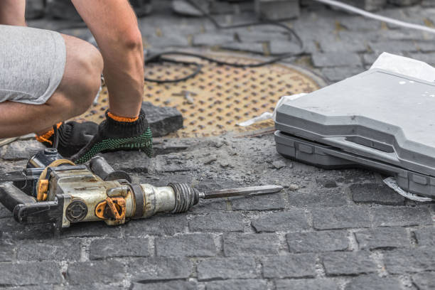 Road worker using a pneumatic drill to break up tarmac around a drain cover on a city street. The man working is wearing high visibility orange workwear and protective work boots. A wheelbarrow and spade is behind him.