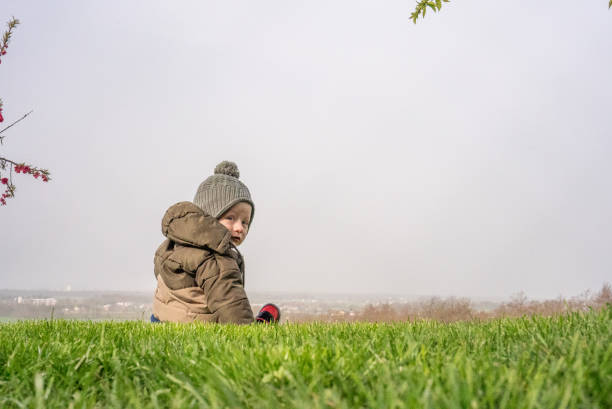 Cute Adorable Baby Boy in Warm Clothing Looking over Shoulder while Sitting on Grassy Field with Clear Sky in the Background during Winter