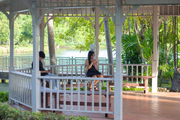 Two young thai adults are sitting for each one in a pavillon in Wachirabenchathat Park (Rot Fai Park) in Bangkok Chatuchak at afternoon..Both are using mobile phone and they are sitting separated in distance. In background is one of lakes in park. Man and woman both have natural black hair. They are looking at screens of phones