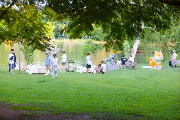 Thai people and young adults are relaxing with pets in  Wachirabenchathat Park (Rot Fai Park) in Bangkok Chatuchak at afternoon. People are in special pet area where dogs can run freely behind small fence around area. People are in groups at several benches. Between people is a pet stroller. In background is a lake. People are wearing casual clothes