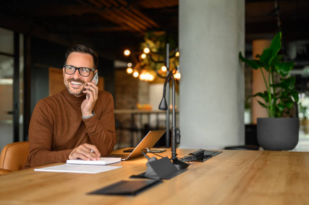 istock Male programmer smiling and discussing project over phone call while working on laptop at desk in office 2170700260