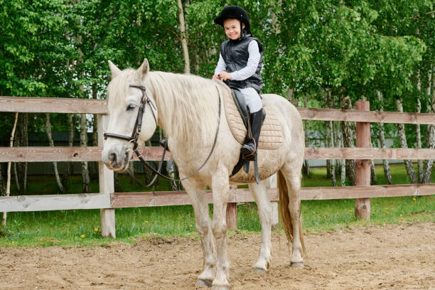 Smiling girl sitting on white horse in paddock surrounded by green trees promoting joy and outdoor activity on a sunny day