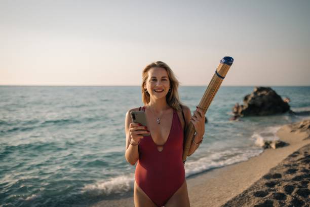 Smiling young woman carrying a beach mat walking and using a smartphone on a sunny summer day at the seaside
