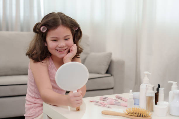 istock Young half-Asian girl smiling while looking in handheld mirror. Hair in soft rollers, surrounded by beauty products on white table. Expressing self-care and happiness during beauty routine at home. 2170715587