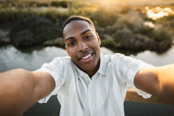 Young man is taking a selfie while smiling by a river at golden hour
